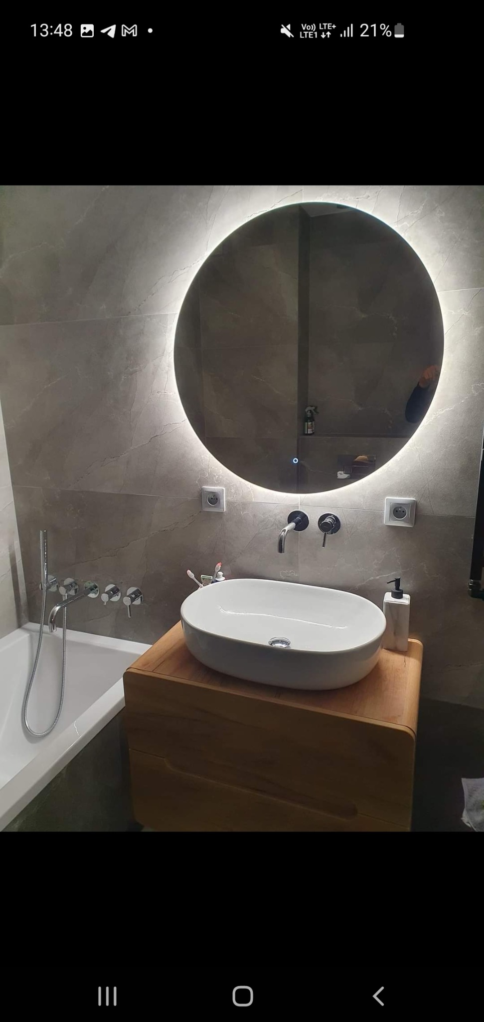 Nowoczesna łazienka z okrągłym lustrem podświetlanym LED, umywalką nablatową na drewnianej szafce i chromowaną armaturą ścienną na tle szarych płytek imitujących marmur.