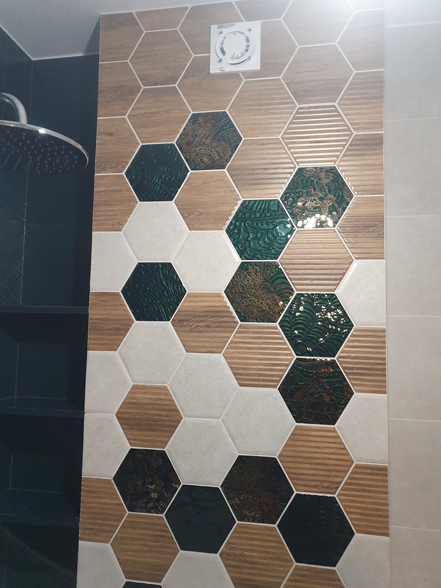 Ściana łazienki wyłożona heksagonalnymi płytkami w odcieniach drewna, bieli i zieleni z wzorami, widoczny fragment deszczownicy i wentylator wyciągowy.