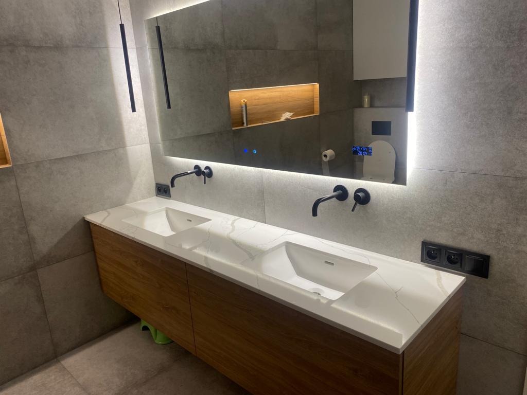 Nowoczesna łazienka z podwójną umywalką na drewnianej szafce z blatem imitującym marmur, czarne baterie, duże lustro z podświetleniem LED i wbudowaną półką, szare płytki na ścianach i podłodze...