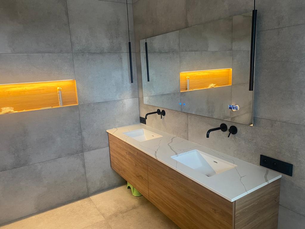 Nowoczesna łazienka z szarymi płytkami, drewnianą szafką pod umywalką z blatem imitującym marmur, czarną armaturą i podświetlaną wnęką w ścianie.