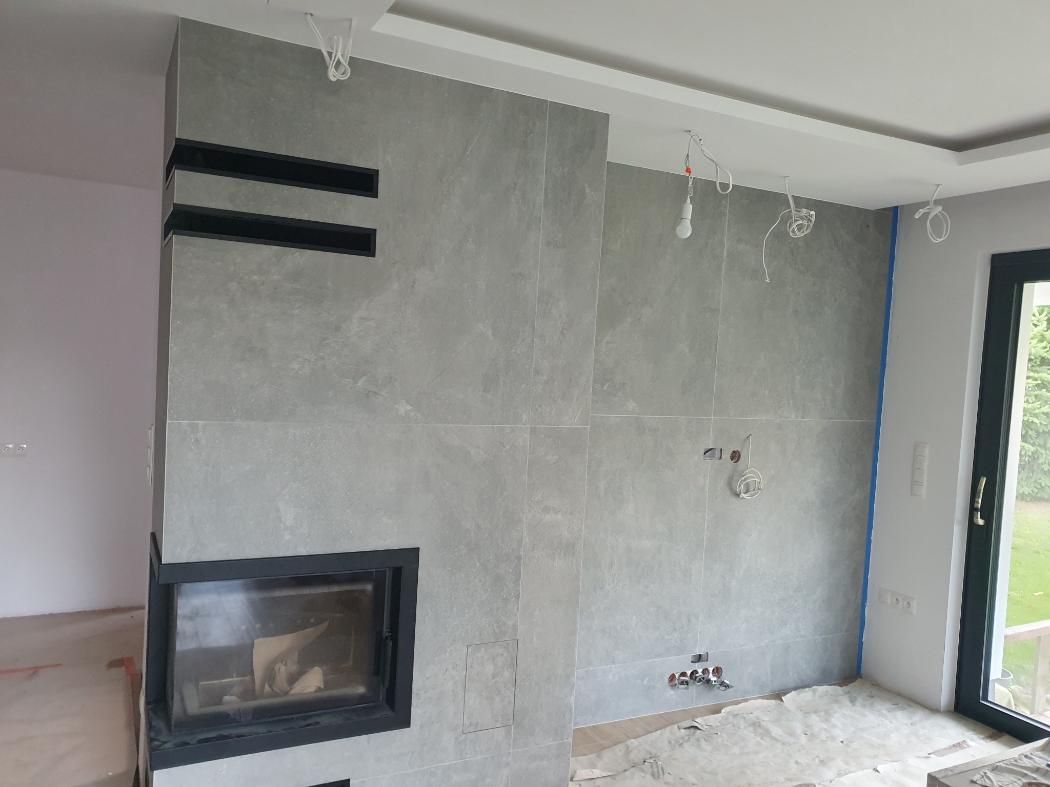 Wnętrze w trakcie prac wykończeniowych: szare płytki imitujące kamień na ścianie z wbudowanym kominkiem z czarną obudową, widoczne przewody elektryczne i instalacja wodna, podłoga zabezpieczona...