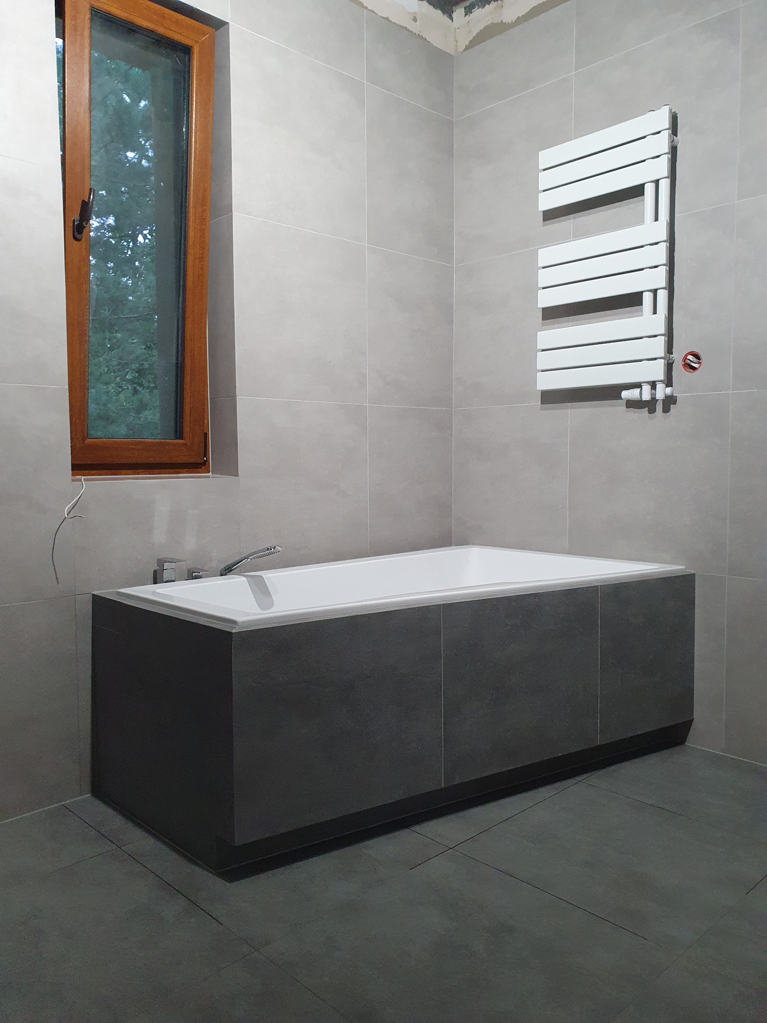 Nowoczesna łazienka w szarościach z wanną obudowaną ciemnymi płytkami, grzejnikiem drabinkowym i oknem z drewnianą ramą; widoczne niedokończone prace przy suficie i wystający kabel.