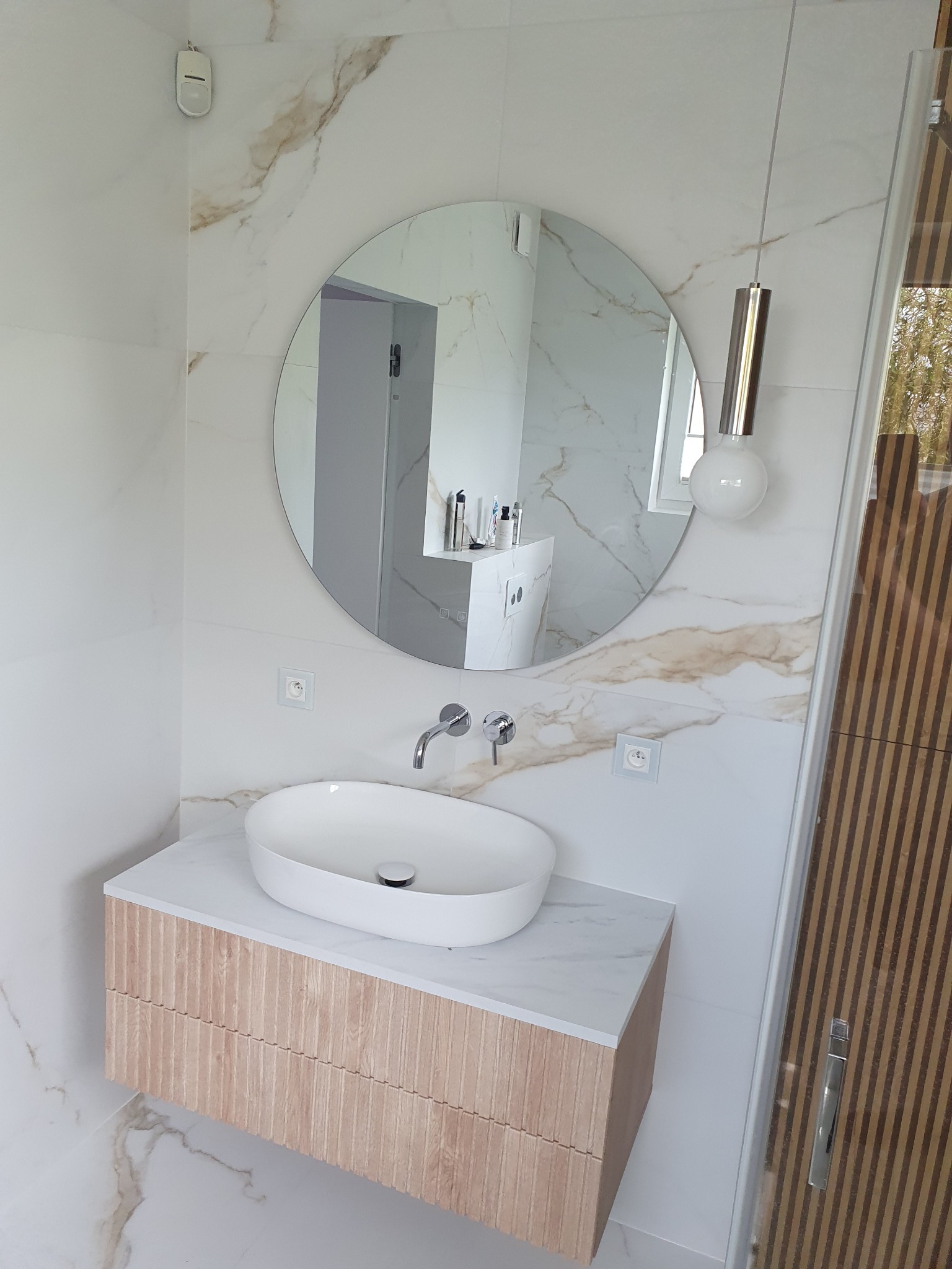 Nowoczesna łazienka z okrągłym lustrem, owalną umywalką nablatową i szafką z ryflowanym frontem, wykończona białymi płytkami imitującymi marmur z delikatnym żyłowaniem.
