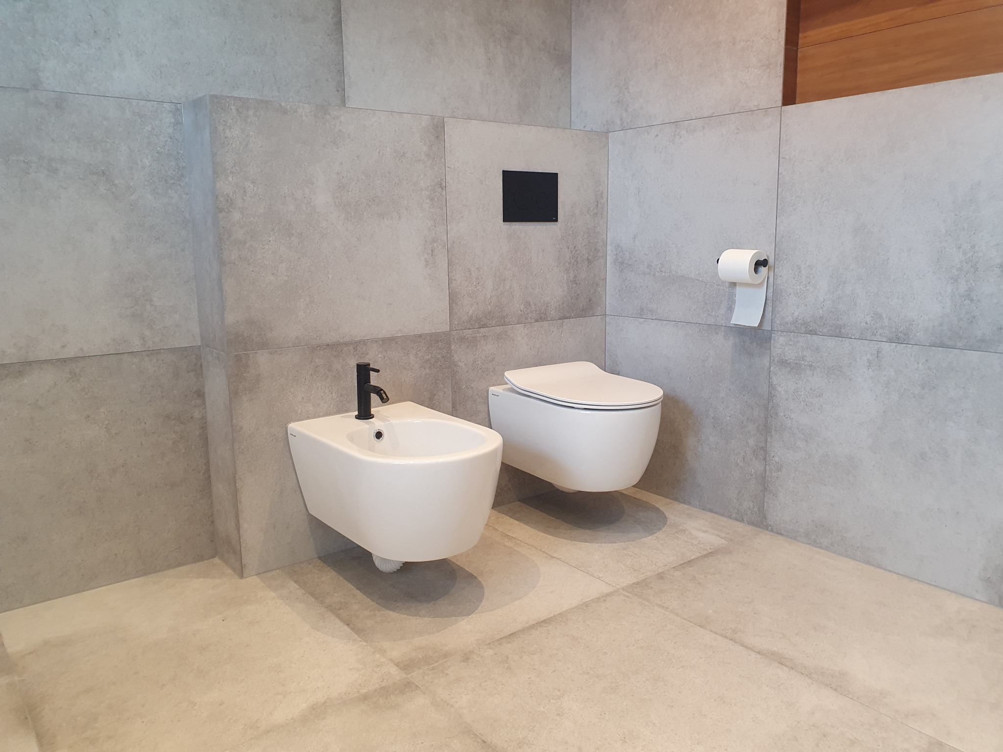 Nowoczesna łazienka z szarymi płytkami, wiszącą miską WC i bidetem z czarną armaturą, minimalistyczny design.