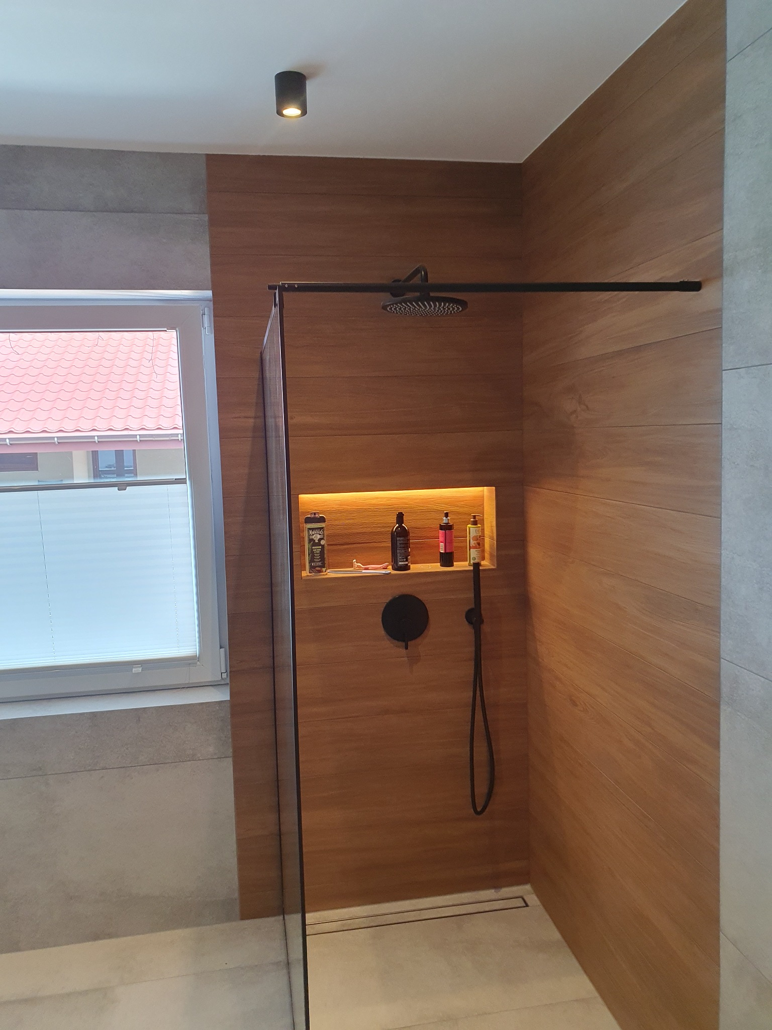 Nowoczesna kabina prysznicowa walk-in z deszczownicą, wykończona płytkami imitującymi drewno, z wnęką podświetloną LED z kosmetykami, widok na okno z żaluzjami plisowanymi.