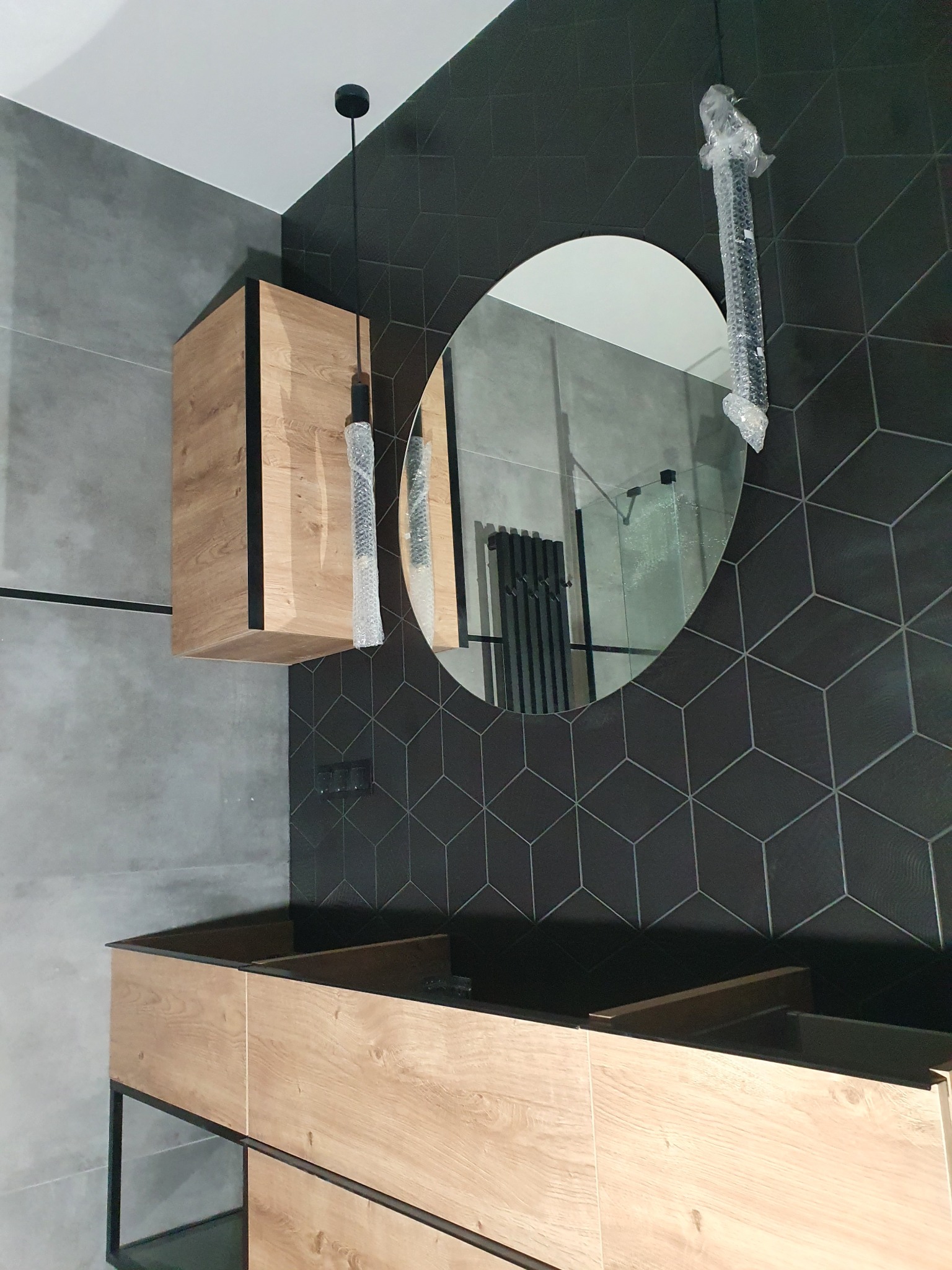 Nowoczesna łazienka z szafkami z jasnego drewna, okrągłym lustrem i czarną, geometryczną mozaiką na ścianie. Elementy oświetlenia w folii ochronnej.