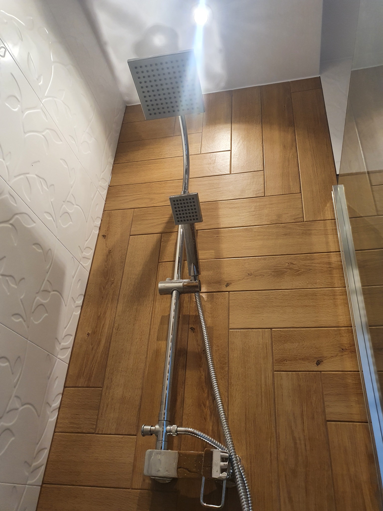 Nowoczesna kabina prysznicowa z deszczownicą i baterią, wykończona płytkami imitującymi drewno ułożonymi w jodełkę, oraz białymi płytkami z delikatnym wzorem roślinnym.