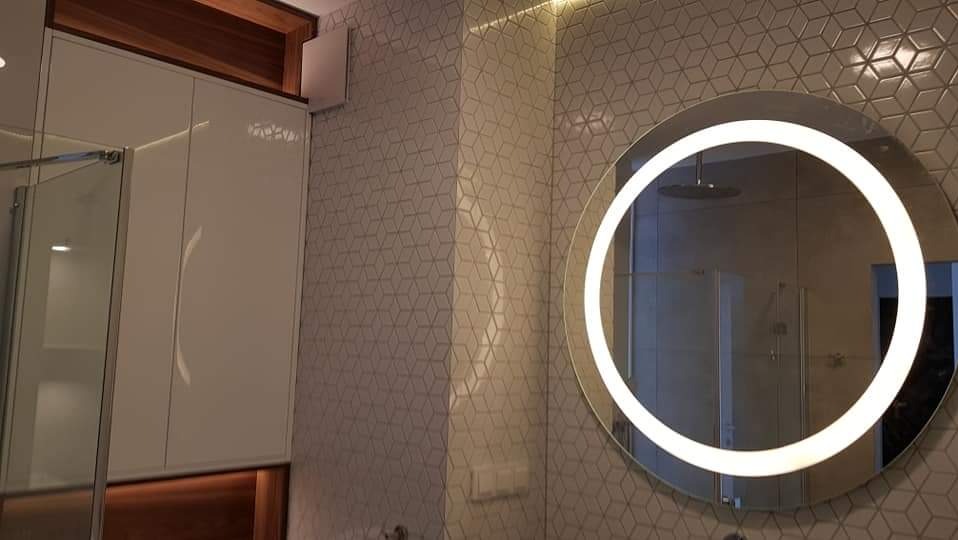 Nowoczesna łazienka z okrągłym lustrem podświetlanym LED, geometryczne płytki na ścianie, białe szafki z chromowanymi uchwytami i widoczna kabina prysznicowa.