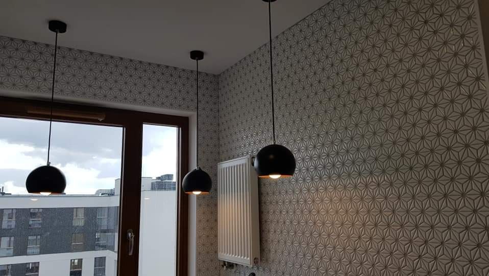 Wnętrze z geometryczną tapetą, trzema czarnymi lampami kulami i grzejnikiem ściennym pod oknem z widokiem na miasto.