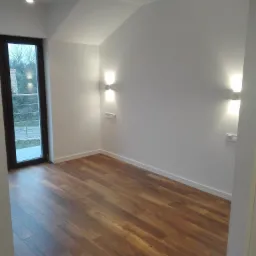 Pusty pokój z brązową podłogą, białymi ścianami, oknem i dwoma kinkietami z gniazdkami, ukazujący minimalistyczny wystrój i gotowość do zamieszkania.