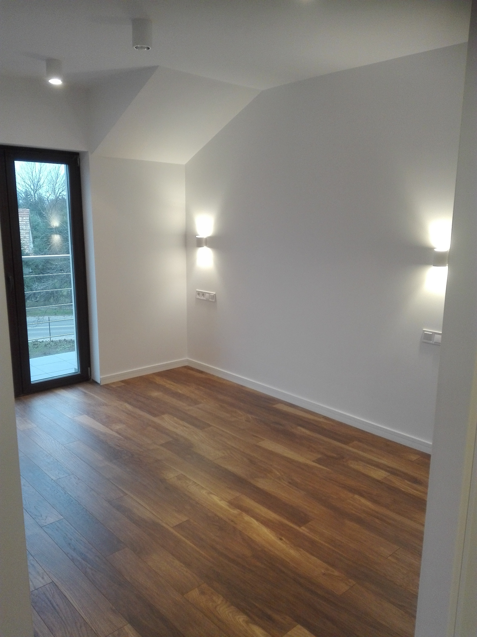 Pusty pokój z brązową podłogą, białymi ścianami, oknem i dwoma kinkietami z gniazdkami, ukazujący minimalistyczny wystrój i gotowość do zamieszkania.