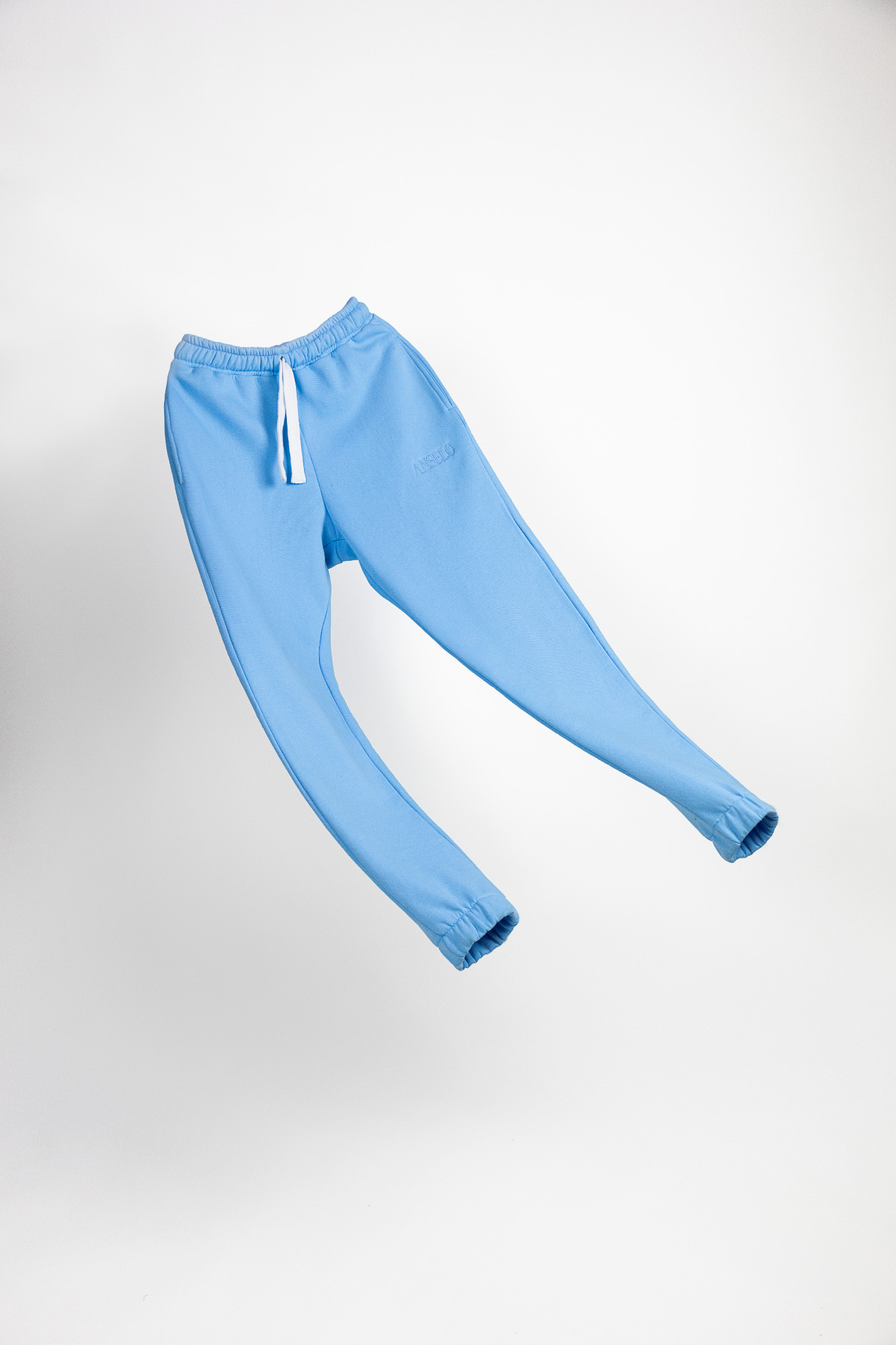 Jasnoniebieskie spodnie dresowe z białym sznurkiem w pasie, unoszące się na białym tle, z delikatnym haftowanym logo w pasie.