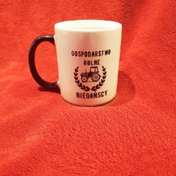 Biały kubek z czarnym uchem na czerwonym tle, z nadrukiem 'Gospodarstwo Rolne Bieganscy' i grafiką traktora w wieńcu.