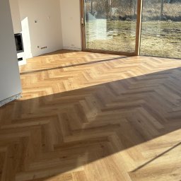 DIR Company Montaż Podłóg - Jasne panele ułożone we wzór jodełki w przestronnym pokoju z dużym oknem. Widoczne detale wykończenia i naturalne światło podkreślające strukturę podłogi.