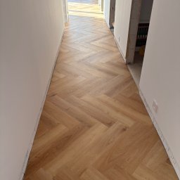 DIR Company Montaż Podłóg - Jasne panele ułożone we wzór jodełki w korytarzu, widok z góry. Świeżo położona podłoga, ściany w trakcie wykańczania. W tle widoczne okno i dalsza część pomieszczenia.