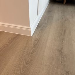 DIR Company Montaż Podłóg - Jasne panele podłogowe ułożone przy białej ścianie z listwą przypodłogową. Widoczny fragment sofy w tle. Gniazdko elektryczne na ścianie.