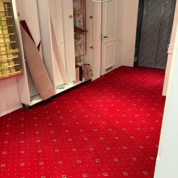 DIR Company Montaż Podłóg - Czerwona wykładzina dywanowa w korytarzu z białymi szafkami i drzwiami. Widoczne piony gazowe. Wykładzina z drobnym, regularnym wzorem w odcieniach złota.