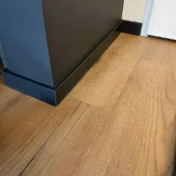 Fragment podłogi z paneli drewnopodobnych, przy krawędzi widoczna czarna listwa przypodłogowa i fragment czarnego mebla. W prawym dolnym rogu stos paneli.