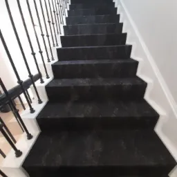 Schody w domu jednorodzinnym, pokryte czarną wykładziną dywanową z delikatnym wzorem, białe stopnice i czarna, metalowa balustrada.