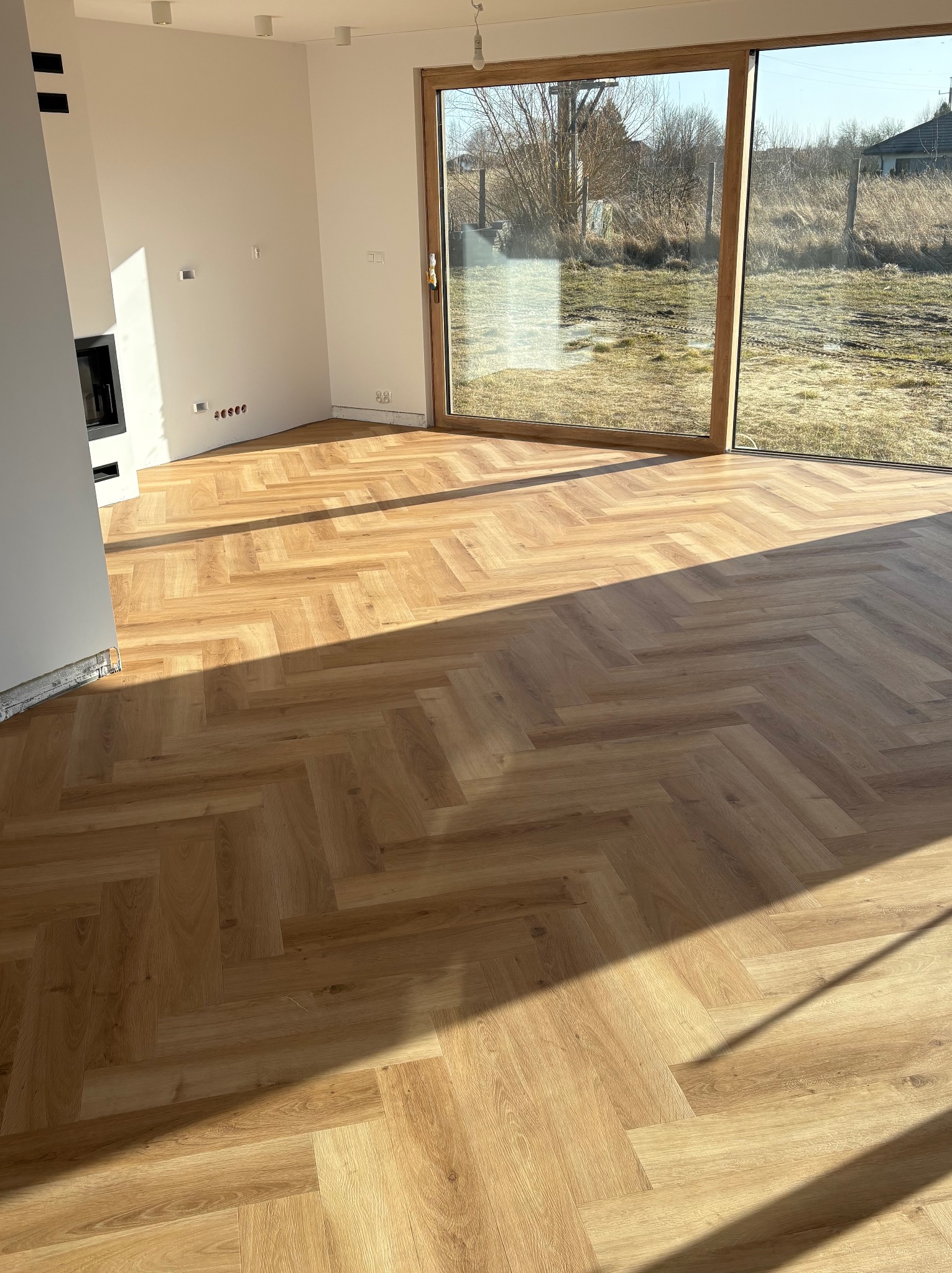 Jasne panele ułożone we wzór jodełki w przestronnym pokoju z dużym oknem. Widoczne detale wykończenia i naturalne światło podkreślające strukturę podłogi.