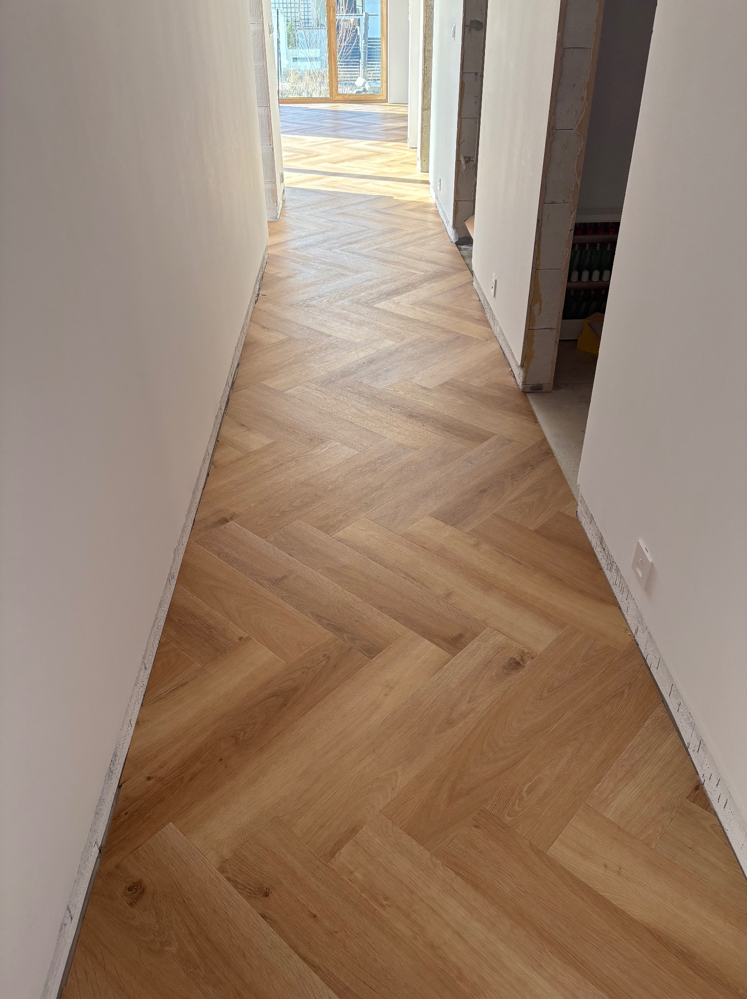 Jasne panele ułożone we wzór jodełki w korytarzu, widok z góry. Świeżo położona podłoga, ściany w trakcie wykańczania. W tle widoczne okno i dalsza część pomieszczenia.