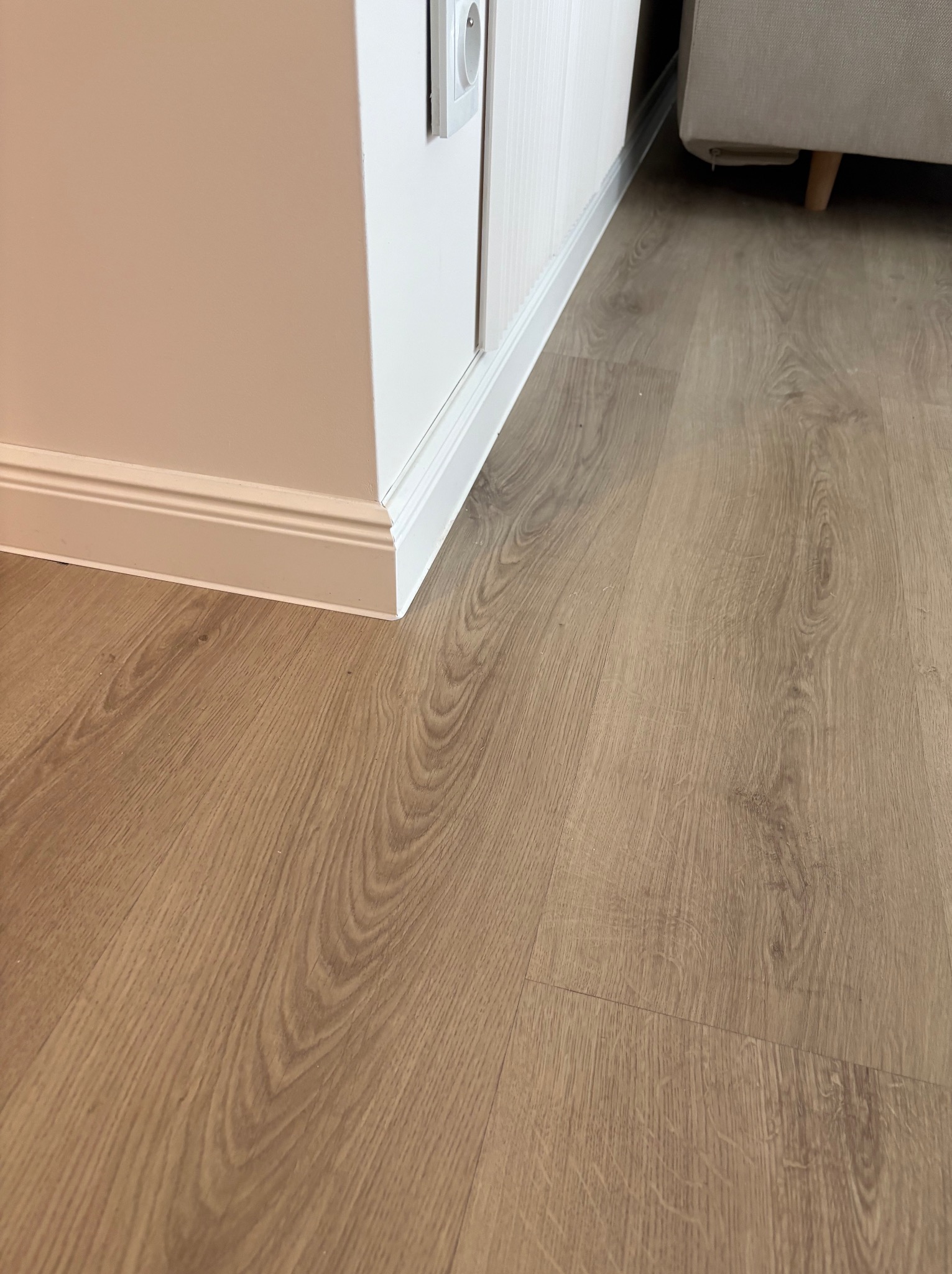 Jasne panele podłogowe ułożone przy białej ścianie z listwą przypodłogową. Widoczny fragment sofy w tle. Gniazdko elektryczne na ścianie.