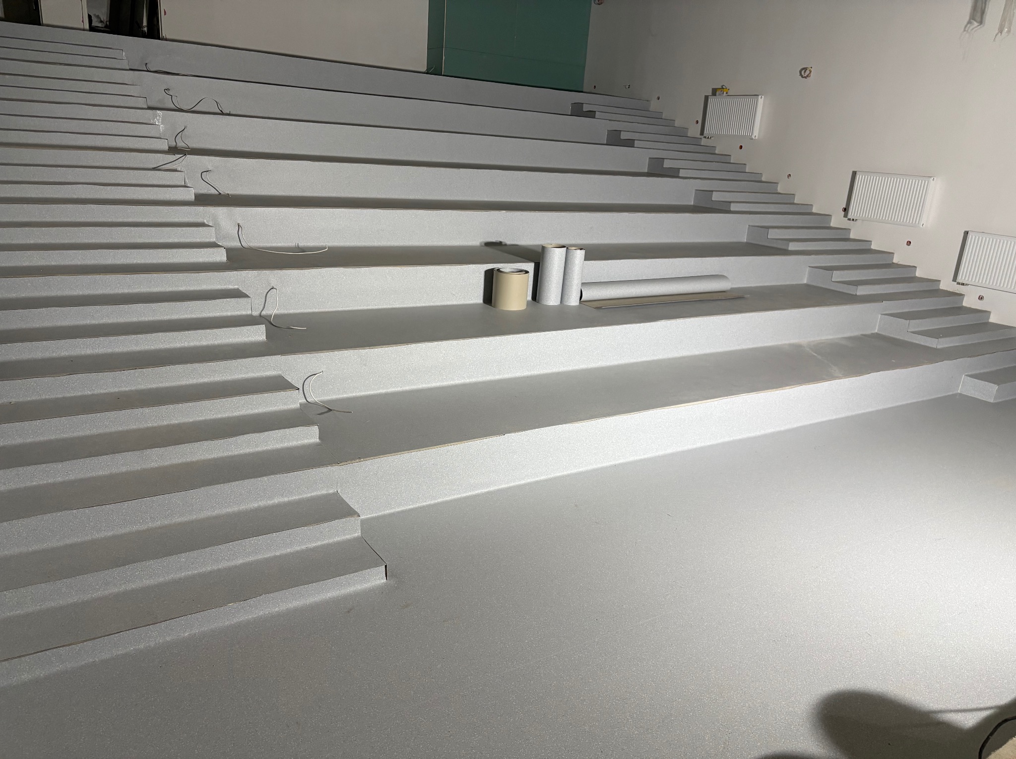 Montaż szarej wykładziny na schodach amfiteatru. Widoczne rolki materiału i taśma. Ściany białe, grzejniki na ścianach. Ujęcie z dołu, szeroki kadr.