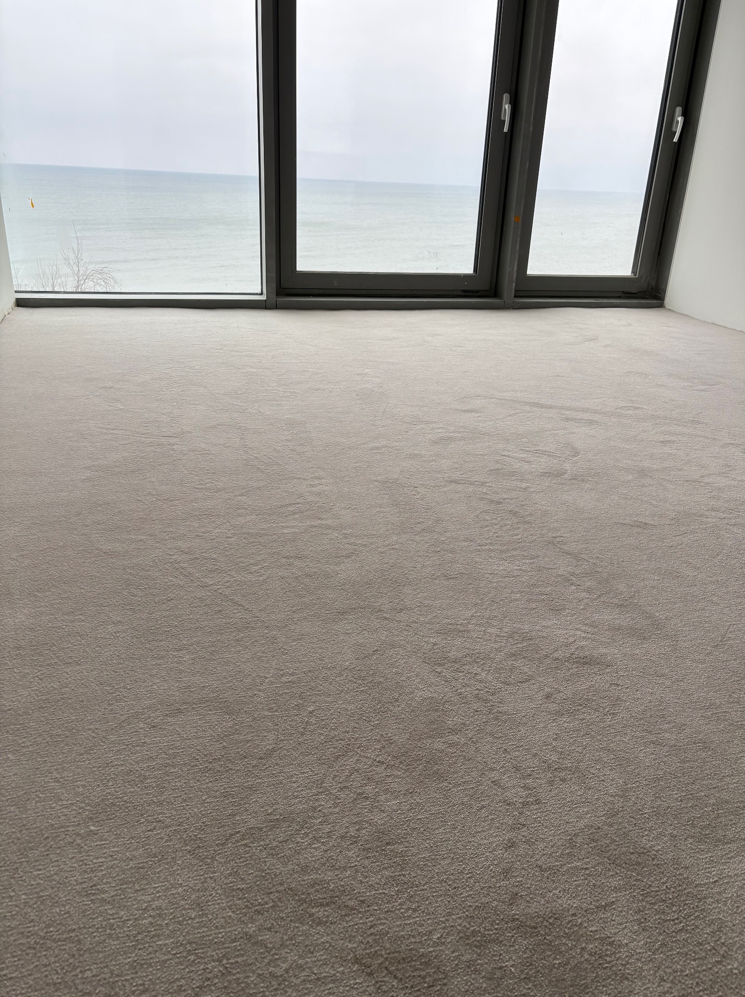 Jasna, beżowa wykładzina dywanowa w pokoju z oknami wychodzącymi na morze. Widok na spokojne wody i pochmurne niebo. Minimalistyczne wnętrze.