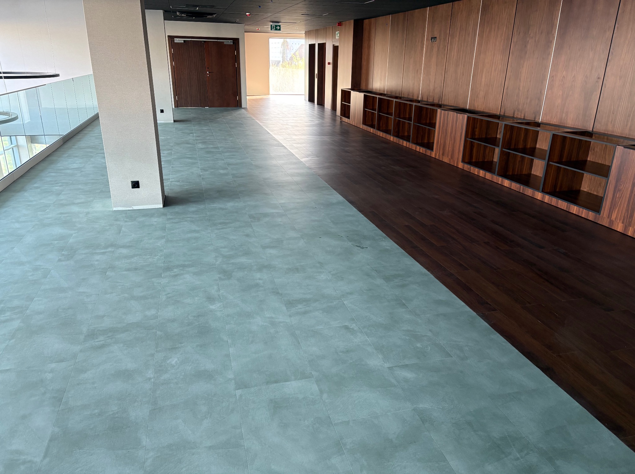 Nowoczesne wnętrze z podziałem podłogi: szara, matowa wykładzina w płytkach i ciemne panele drewniane. W tle ściana z drewnianymi szafkami i jasne drzwi.