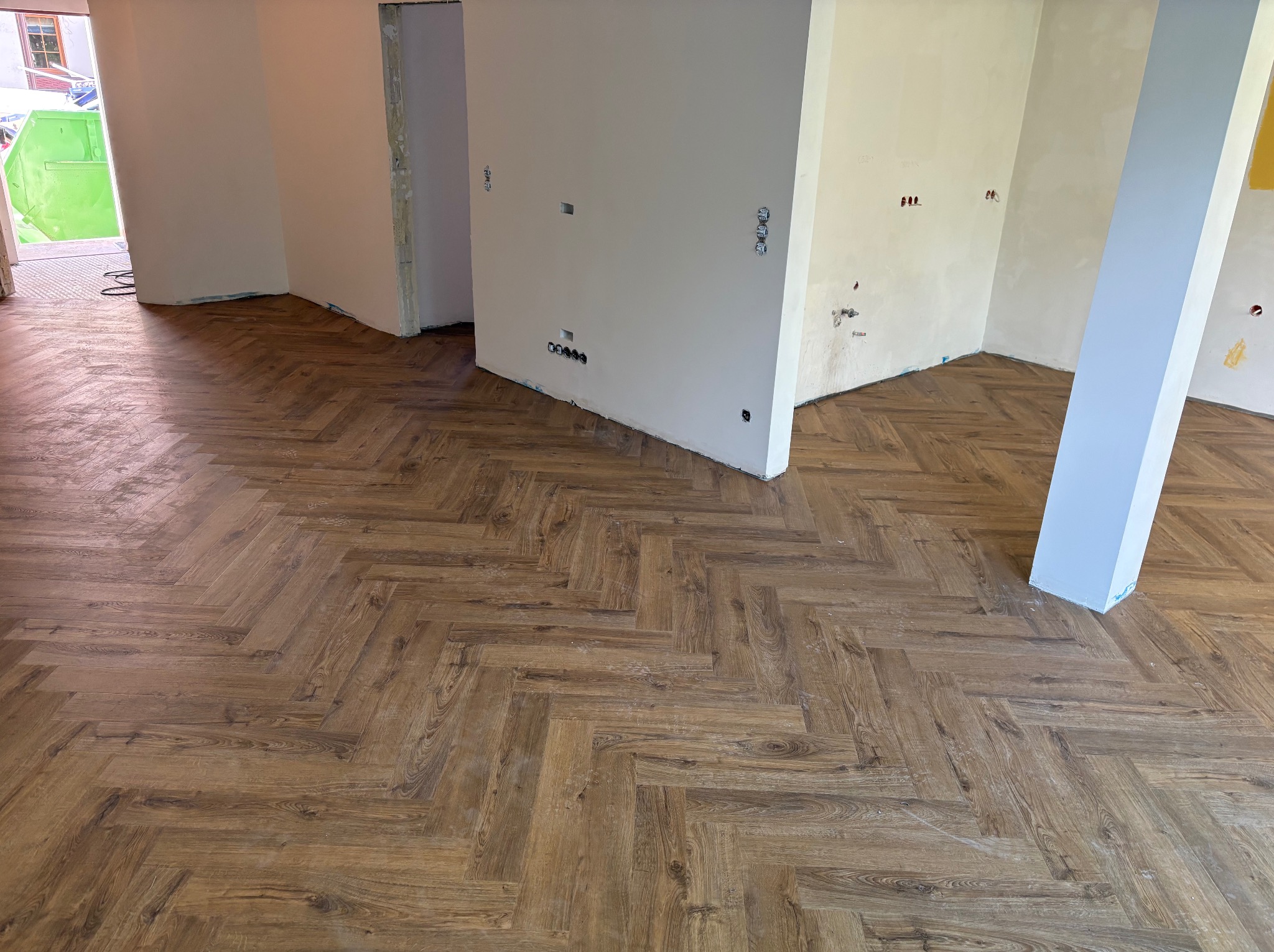 Ukończona podłoga z paneli ułożonych w jodełkę w jasnym wnętrzu. Widoczne gniazdka elektryczne na ścianach i ślady po remoncie. Na zewnątrz kontener budowlany.