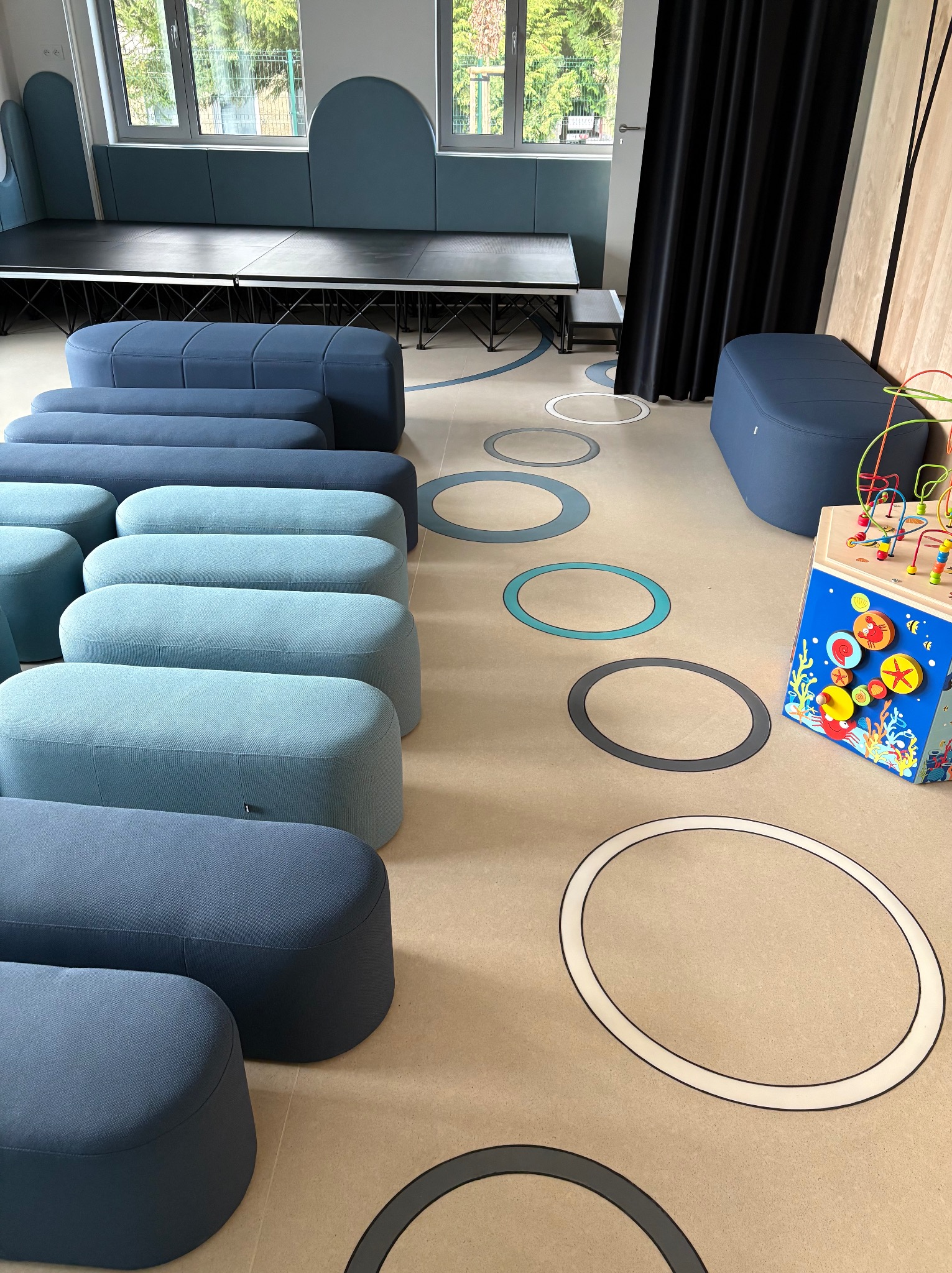 Jasna podłoga z okrągłymi wzorami w odcieniach szarości i błękitu, dopasowane pufy w rzędach i zabawki w pokoju zabaw. Scena z podestem w tle.