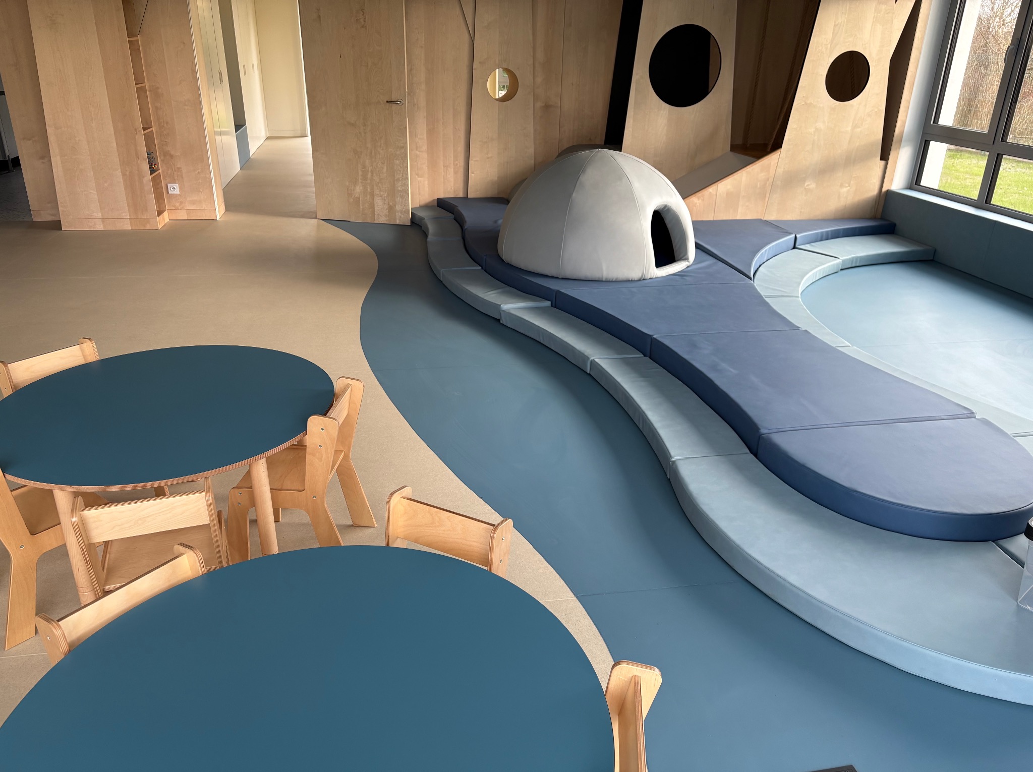 Nowoczesne wnętrze bawialni z podłogą w odcieniach błękitu i beżu, drewnianymi meblami oraz strefą relaksu z miękkimi siedziskami i igloo. Jasne, naturalne światło.