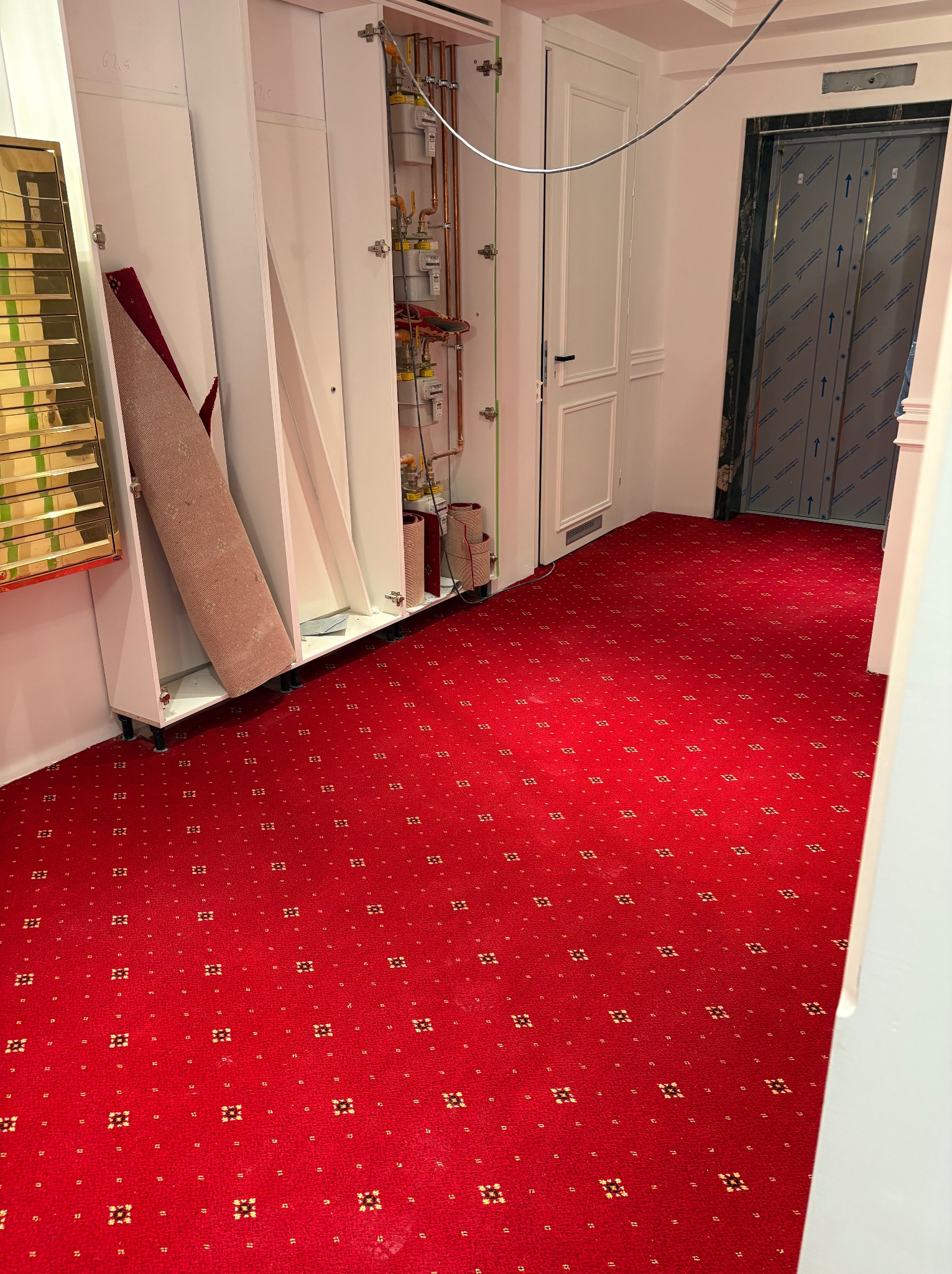 Czerwona wykładzina dywanowa w korytarzu z białymi szafkami i drzwiami. Widoczne piony gazowe. Wykładzina z drobnym, regularnym wzorem w odcieniach złota.
