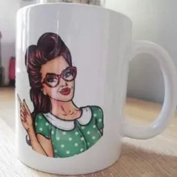 Biały kubek z nadrukiem w stylu pop-art przedstawiającym kobietę w okularach i zielonej sukience w białe kropki, stojący na drewnianym blacie.
