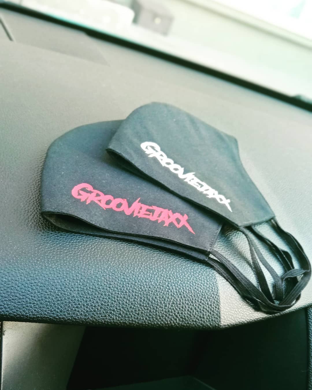 Dwie czarne maseczki ochronne z nadrukowanym logo 'Groovietaxx' w kolorze białym i różowym, leżące na czarnej, teksturowanej powierzchni deski rozdzielczej samochodu.