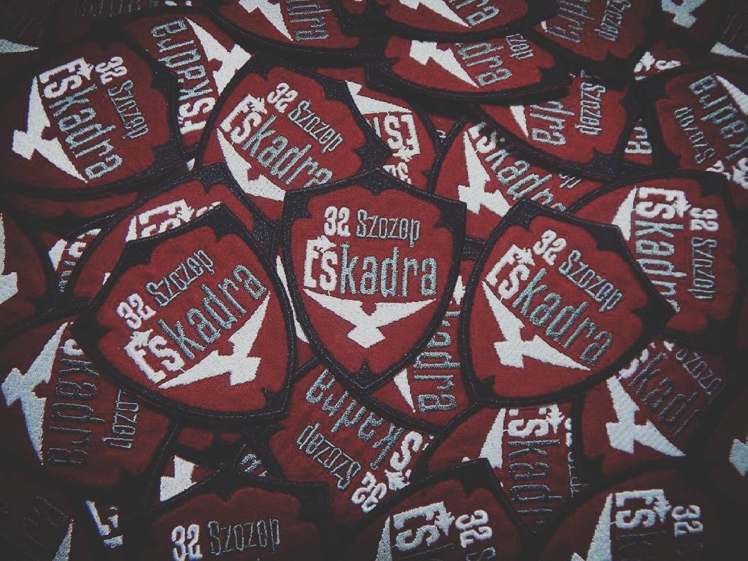Stos naszywek w kształcie tarczy z napisem '32 Szozop Eskadra' i symbolem błyskawicy, wykonanych techniką haftu na czerwonym tle.