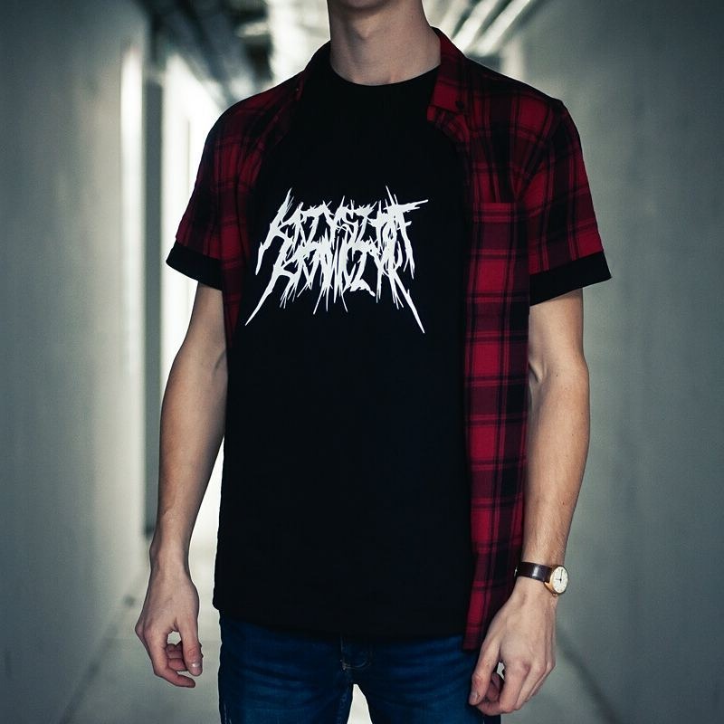 Mężczyzna w czarnej koszulce z białym, ostrym, stylizowanym napisem, w rozpiętej czerwono-czarnej koszuli w kratę, stojący w szarym korytarzu.