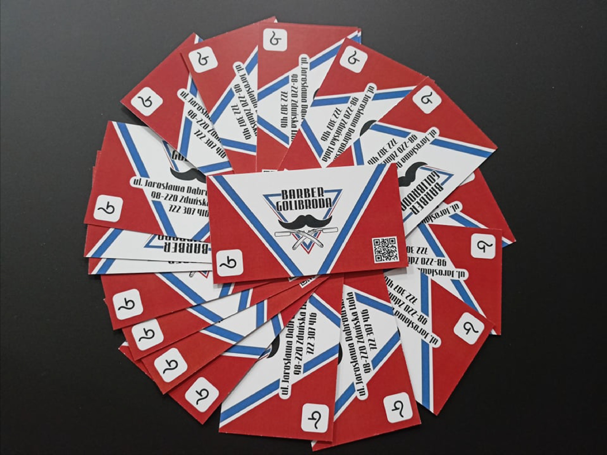 Stos wizytówek z czerwono-biało-niebieskim motywem barberskim, ułożonych w okrąg na czarnym tle, z widocznym adresem i kodem QR.