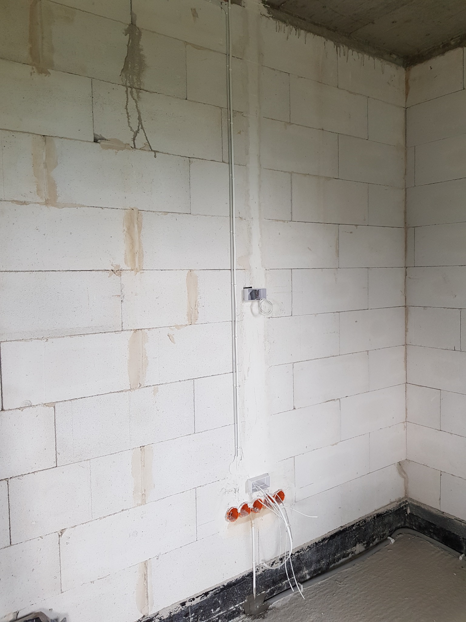 Surowa ściana z betonu komórkowego z widocznymi przewodami elektrycznymi wychodzącymi z pomarańczowych puszek instalacyjnych oraz metalową rurką instalacyjną przymocowaną do ściany.