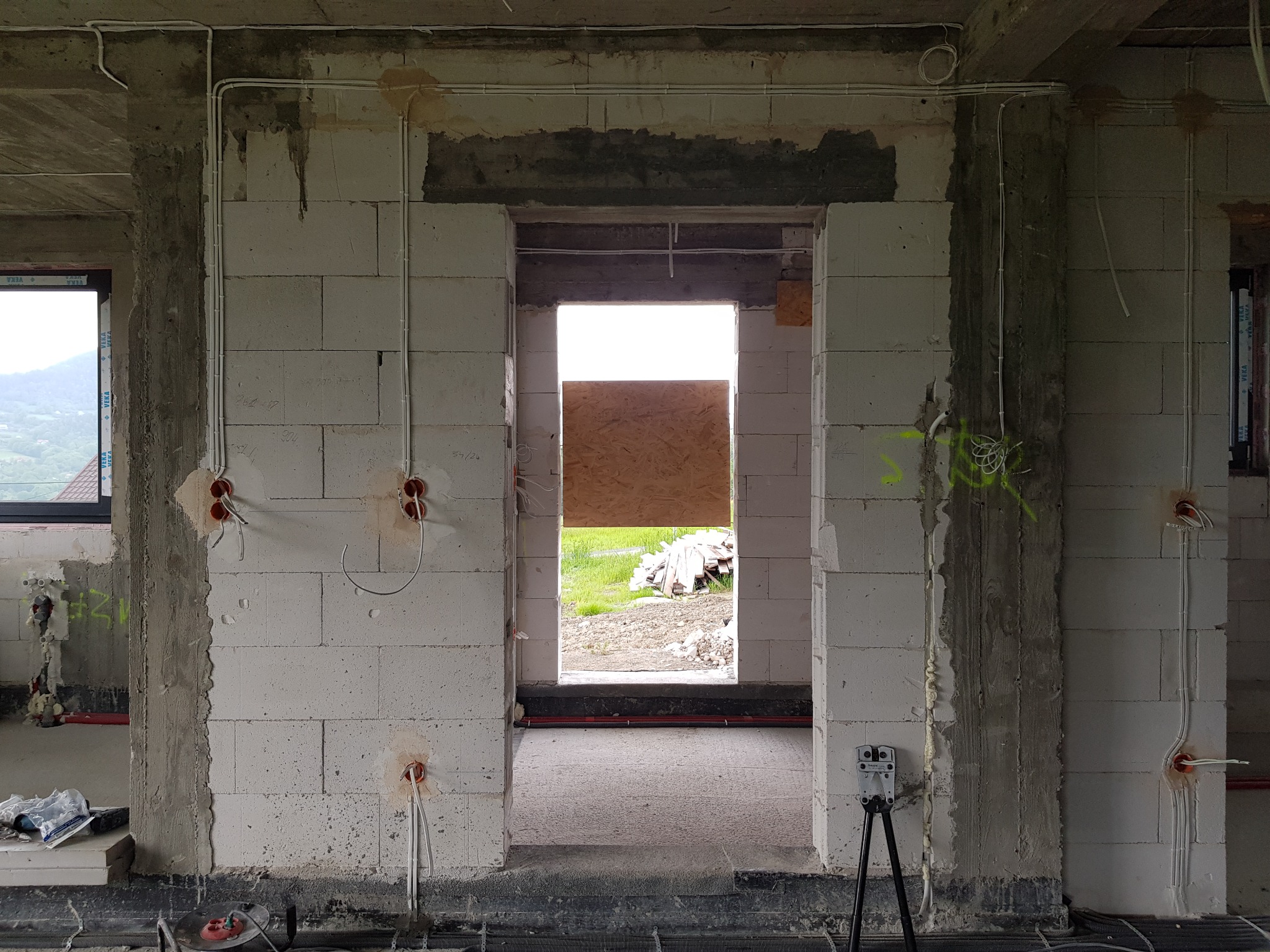 Wewnętrzne ściany budynku w trakcie budowy z widoczną instalacją elektryczną w peszlach, wystające przewody z puszek, widok na zewnątrz przez otwór drzwiowy zasłonięty płytą OSB.