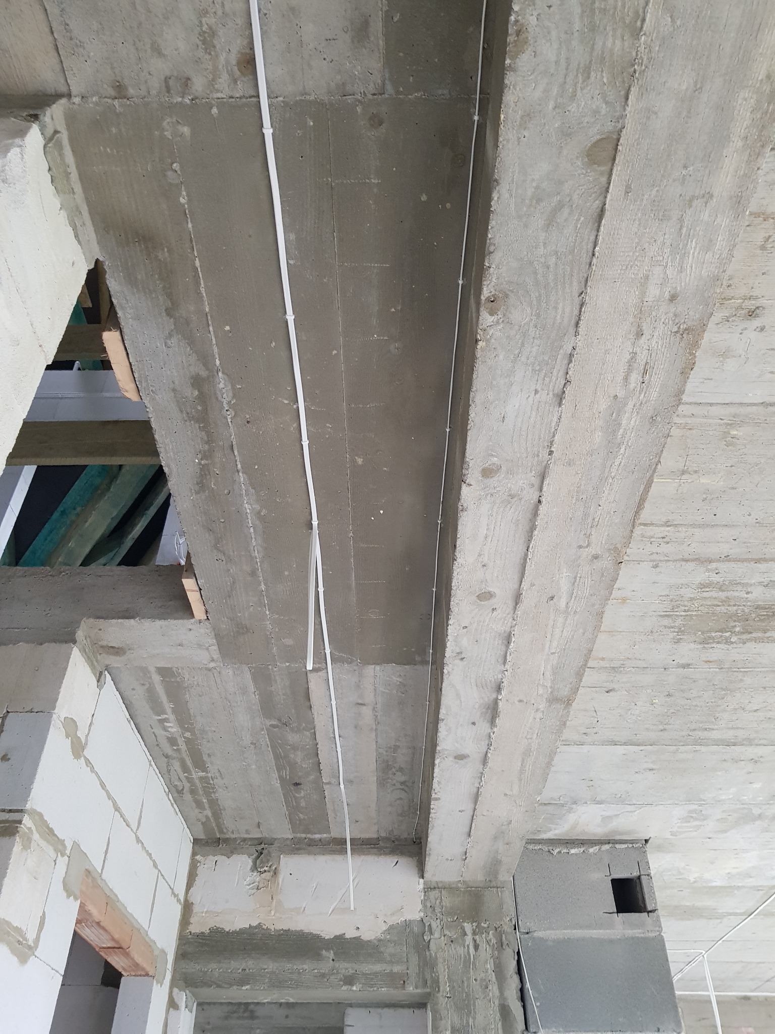 Surowy betonowy strop z widocznymi elementami konstrukcyjnymi i pojedynczym przewodem elektrycznym w białej listwie, zamocowanym pionowo do powierzchni betonu. Widoczne bloczki betonowe i fragmenty...