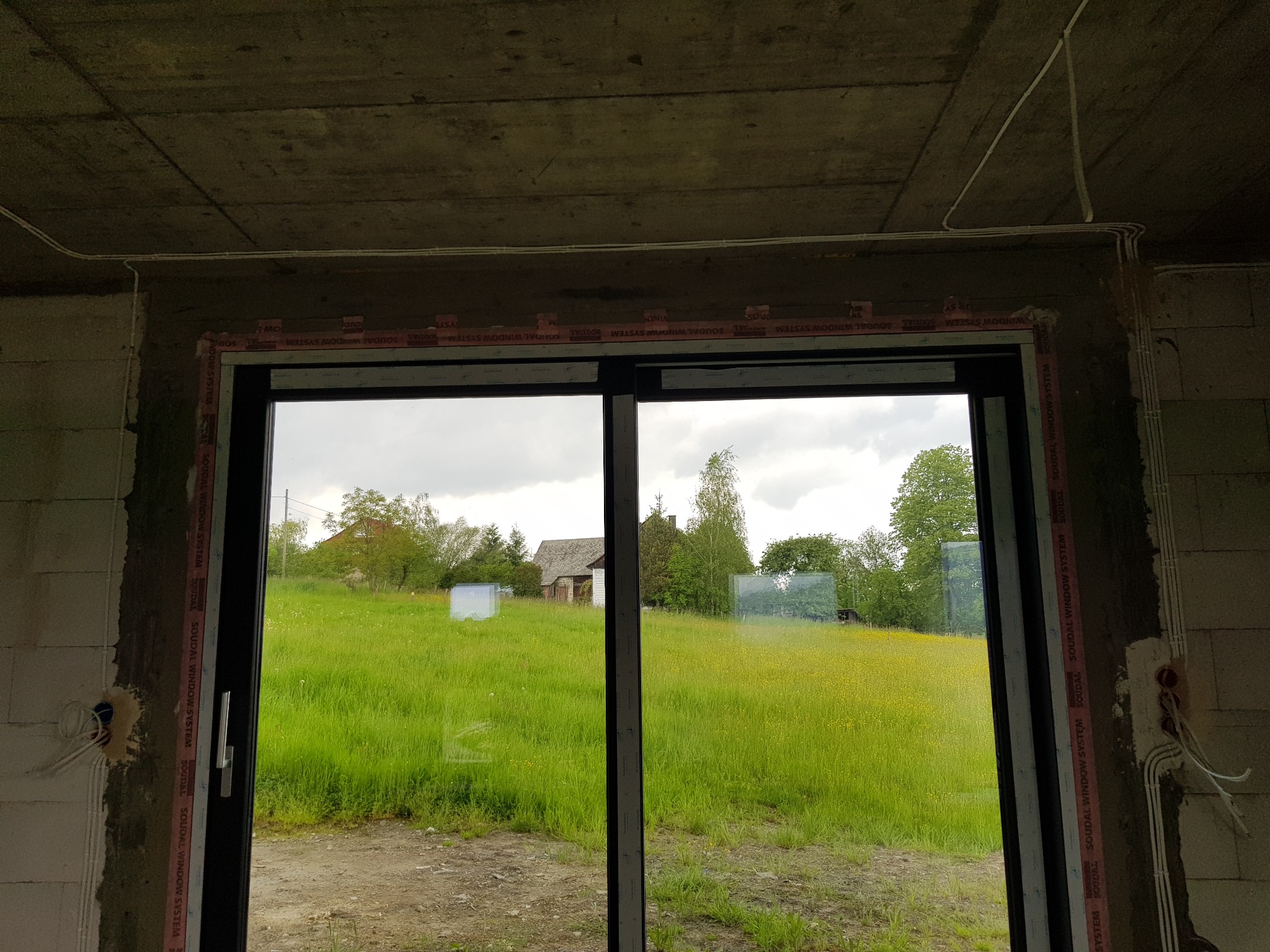 Surowy stan wykończenia wnętrza z widocznymi przewodami elektrycznymi ułożonymi na suficie i ścianach, okno z widokiem na zieloną łąkę i zabudowania.