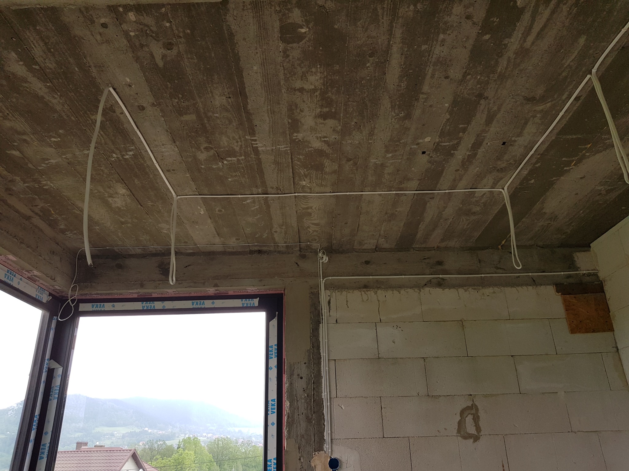 Surowy stan budynku w trakcie prac: widoczne kable elektryczne prowadzone po betonowym suficie i ścianach z bloczków, okno z widokiem na góry w tle.