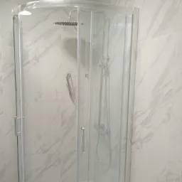 Kabina prysznicowa z deszczownicą i półką na kosmetyki, wykończona białymi płytkami imitującymi marmur.