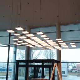 REVOLTA Przemysław Burkowski - Sufit z kwadratowymi panelami LED w nowoczesnym biurze, widoczne przygotowania do remontu, zabezpieczona podłoga płytami OSB, drabina oparta o framugę drzwi, duże okna z widokiem na parking.