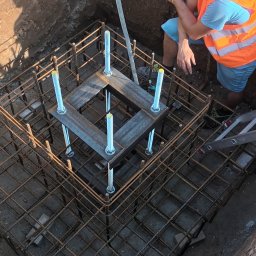 REVOLTA Przemysław Burkowski - Pracownik w pomarańczowej kamizelce i kasku nad wykopem z konstrukcją zbrojeniową i metalowymi elementami mocującymi, widoczne narzędzia elektryczne w tle.