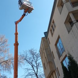 REVOLTA Przemysław Burkowski - Pomarańczowy podnośnik koszowy z pracownikiem na wysokości przy budynku mieszkalnym, błękitne niebo w tle.