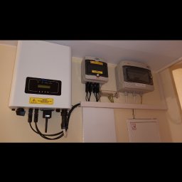 REVOLTA Przemysław Burkowski - Biały inwerter fotowoltaiczny Devo z podłączonymi kablami, obok dwie szare skrzynki elektryczne na ścianie wewnątrz budynku.