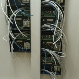 REVOLTA Przemysław Burkowski - Dwa rzędy czarnych skrzynek z elektroniką i licznymi kablami (białe, niebieskie, żółte) wychodzącymi z nich, zamontowane na białej ścianie.