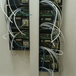 Dwa rzędy czarnych skrzynek z elektroniką i licznymi kablami (białe, niebieskie, żółte) wychodzącymi z nich, zamontowane na białej ścianie.